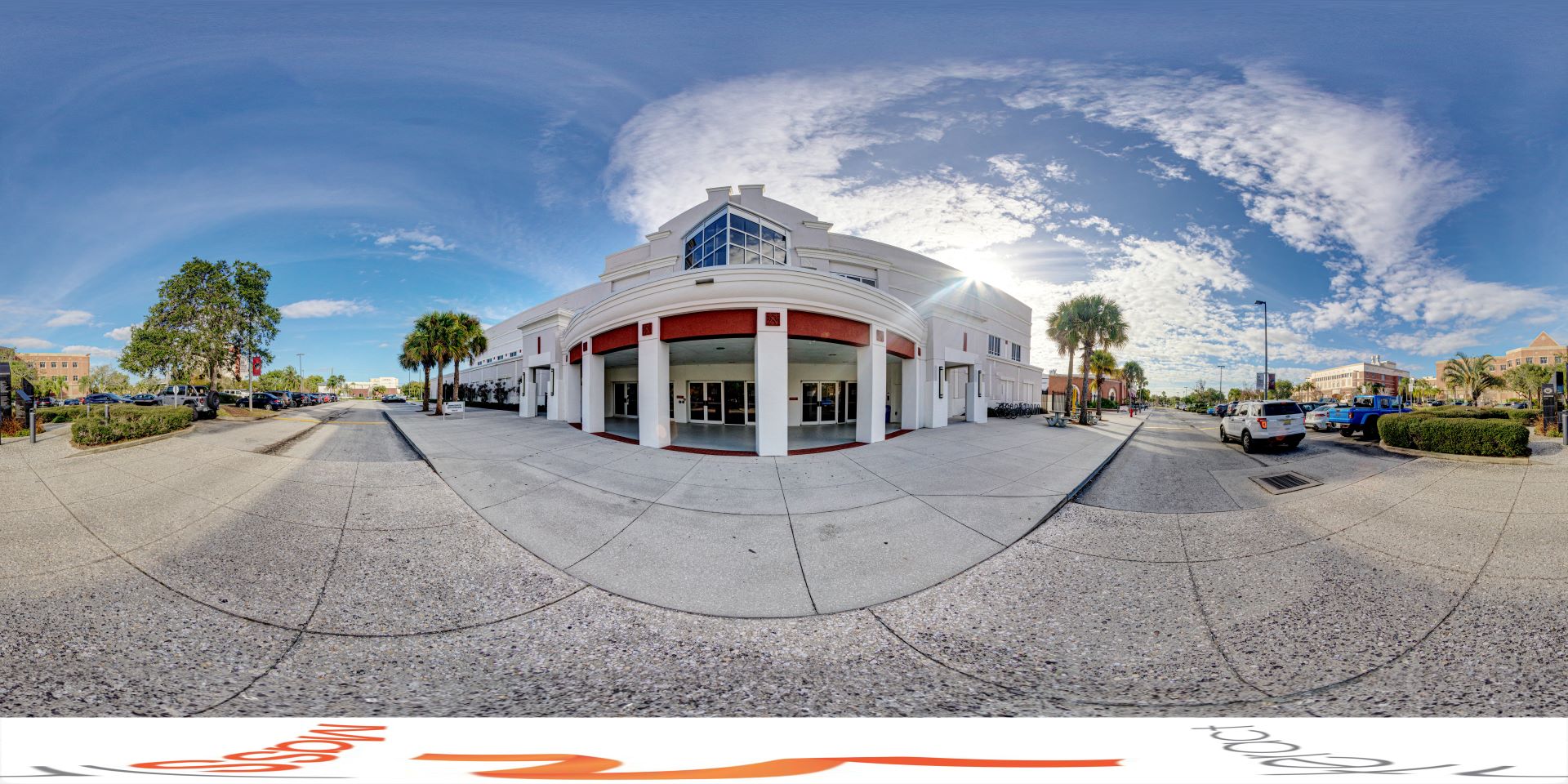 A panoramic view of the Clemente Center at Florida Tech, showcasing the building's exterior with a round entrance, palm trees, parked cars, and a clear blue sky with scattered clouds